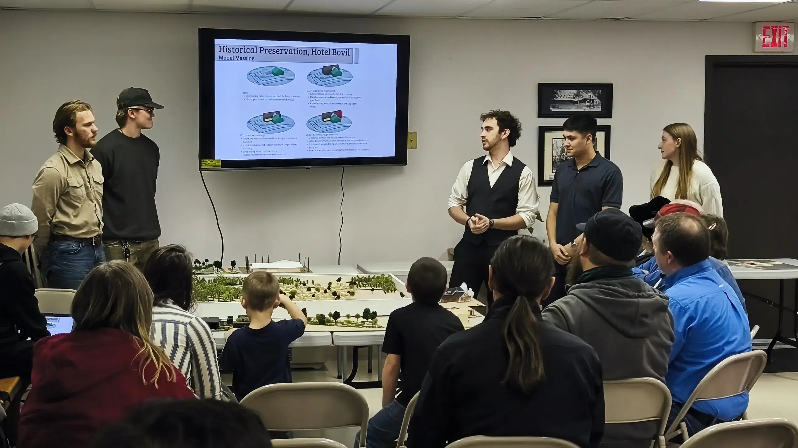 Four men and one woman present in front of a large screen in front of an audience in a classroom.