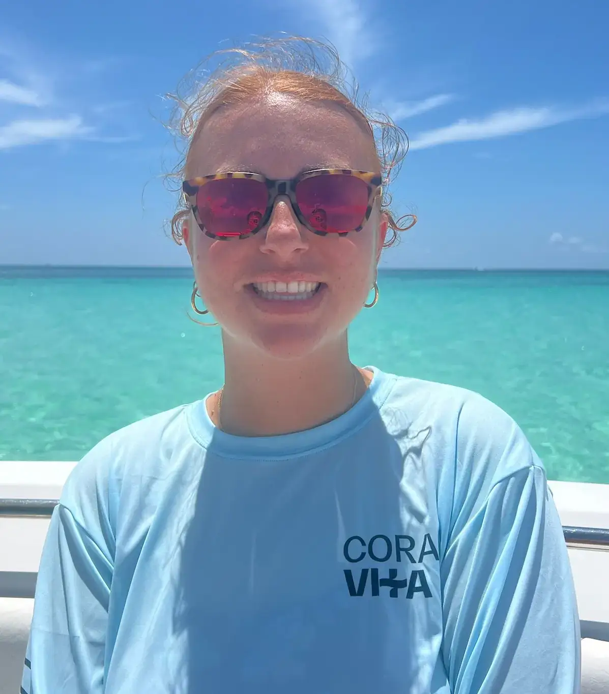 Woman with shades stands with her back to a blue sea.