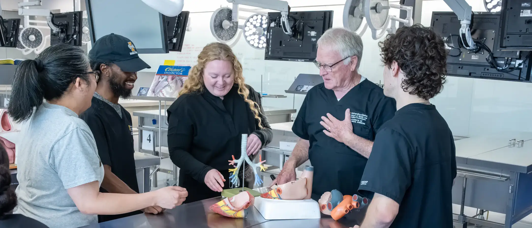 A group of individuals in a laboratory discussing medical models on a table, including anatomical representations of the human body.