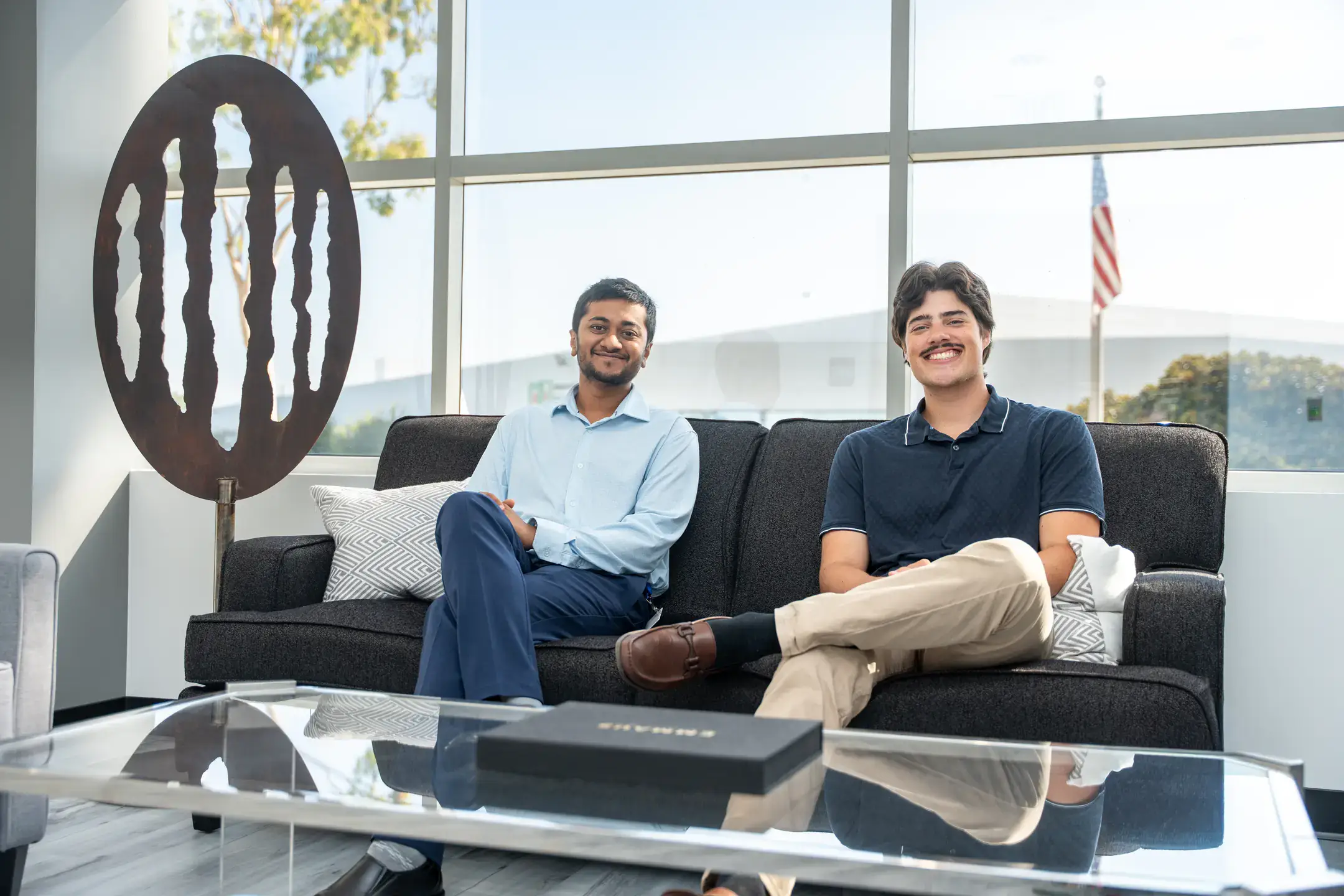 Two men sitting on a sofa in a modern office with a large window behind them, featuring the U.S. flag and outdoor greenery.