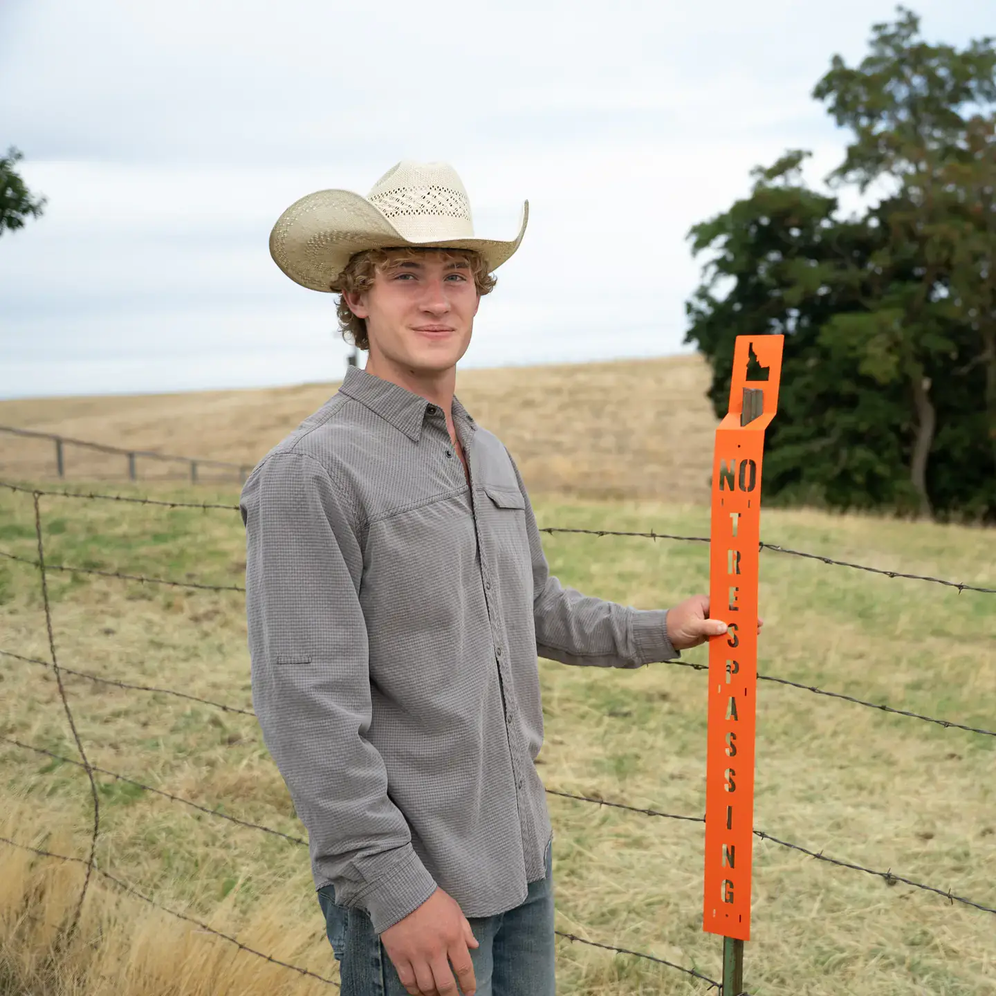 Young man in a cowboy hat holds an orange no trespassing sign by a barbed-wire fence in a rural field.