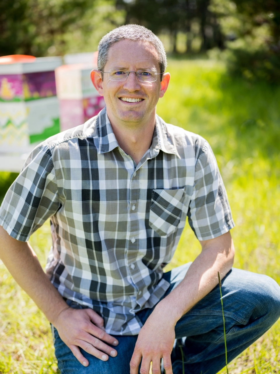 Man in short sleeve shirt kneels in grass in front of beehive.
