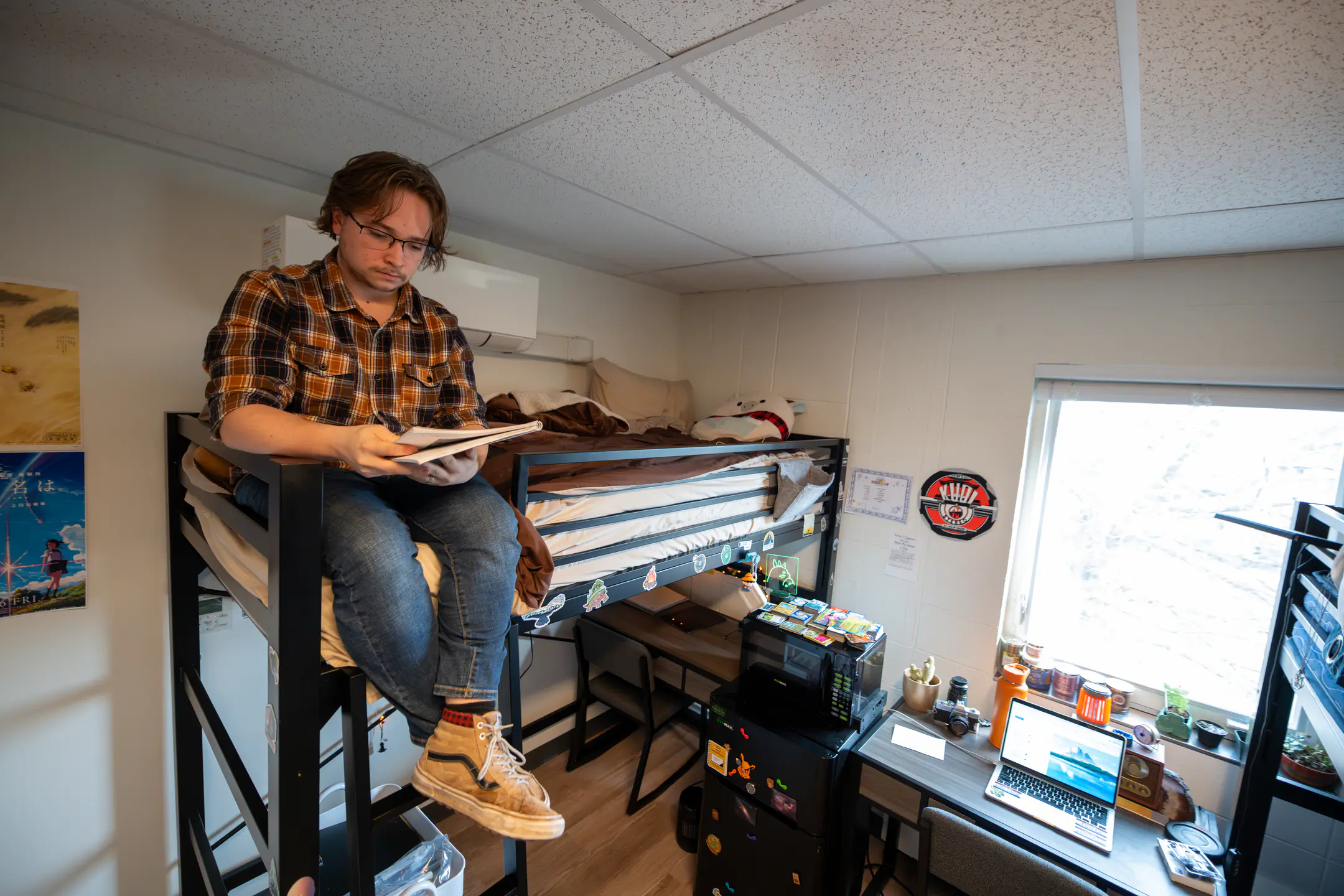 A male student reads from a stack of papers while sitting on the edge of his loft bed.