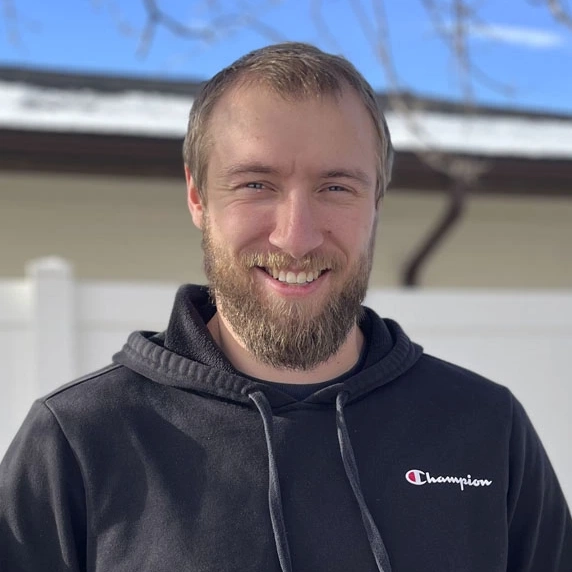 A man smiles for a portrait while standing outdoors with melting snow in the background.