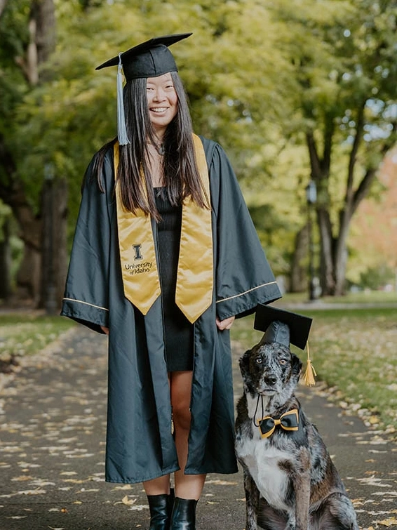 A smiling woman in graduation regalia stands next to her sitting dog, both wearing graduation caps.