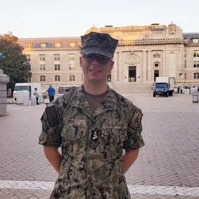 Person in U.S. Navy camouflage uniform standing in a plaza before a large stone building.