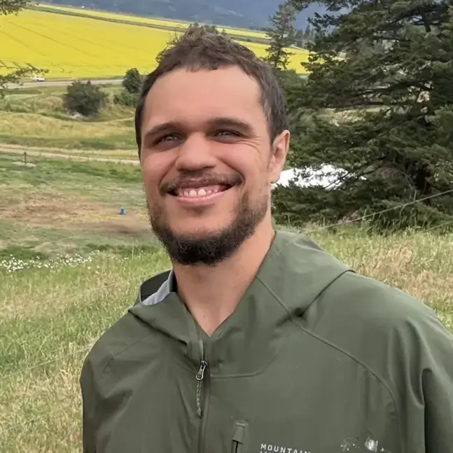 Three people stand outdoors in a grassy field with mountains and yellow flowers visible in the distance.