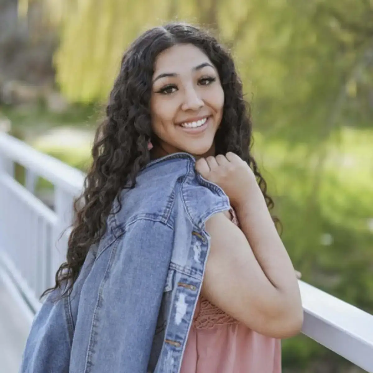 Smiling woman with curly hair holding a denim jacket over her shoulder, standing on a bridge with trees in the background.