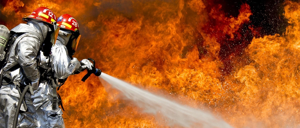 two firefighters dressed in silver heat resistance protective equipment trying to put out part of a large blaze