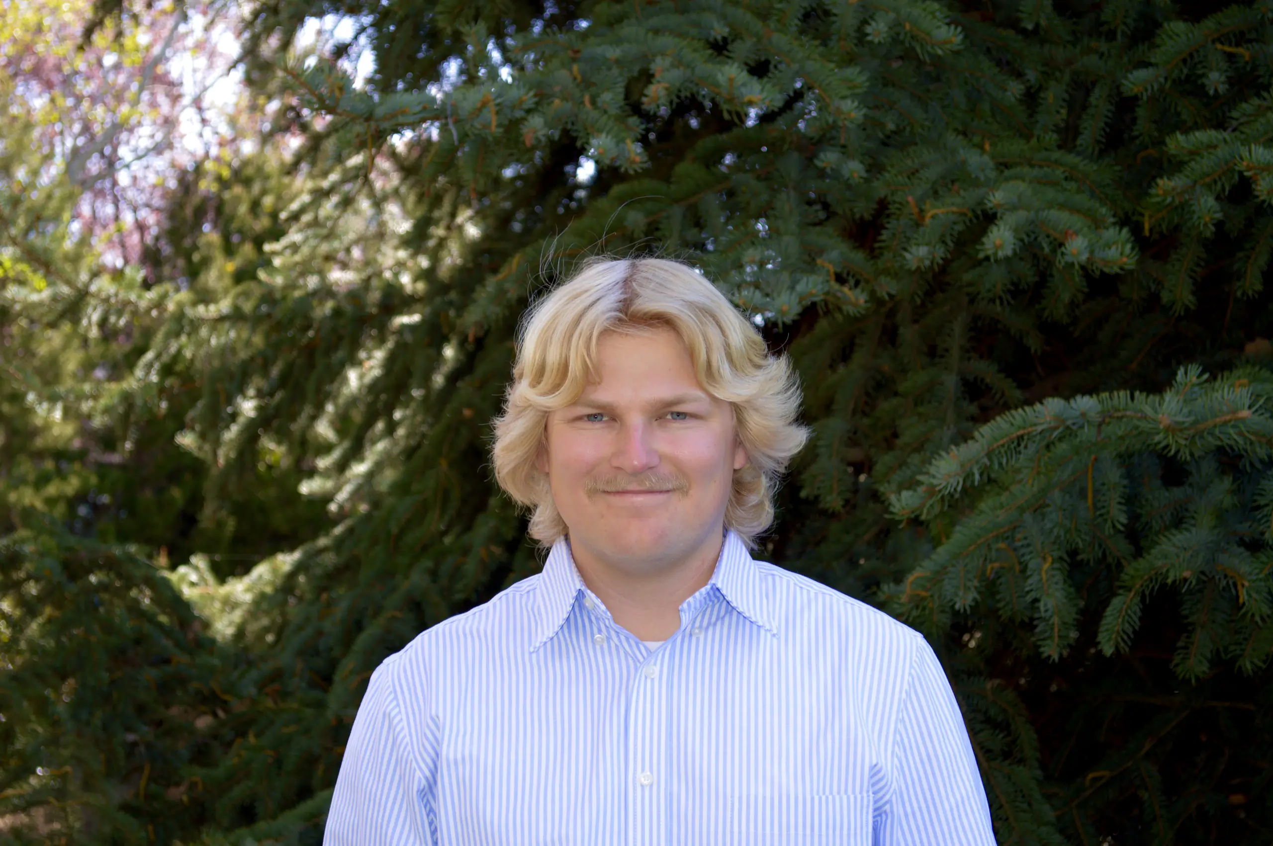 Alex Call smiles wearing a collared shirt in front of evergreen trees in background.