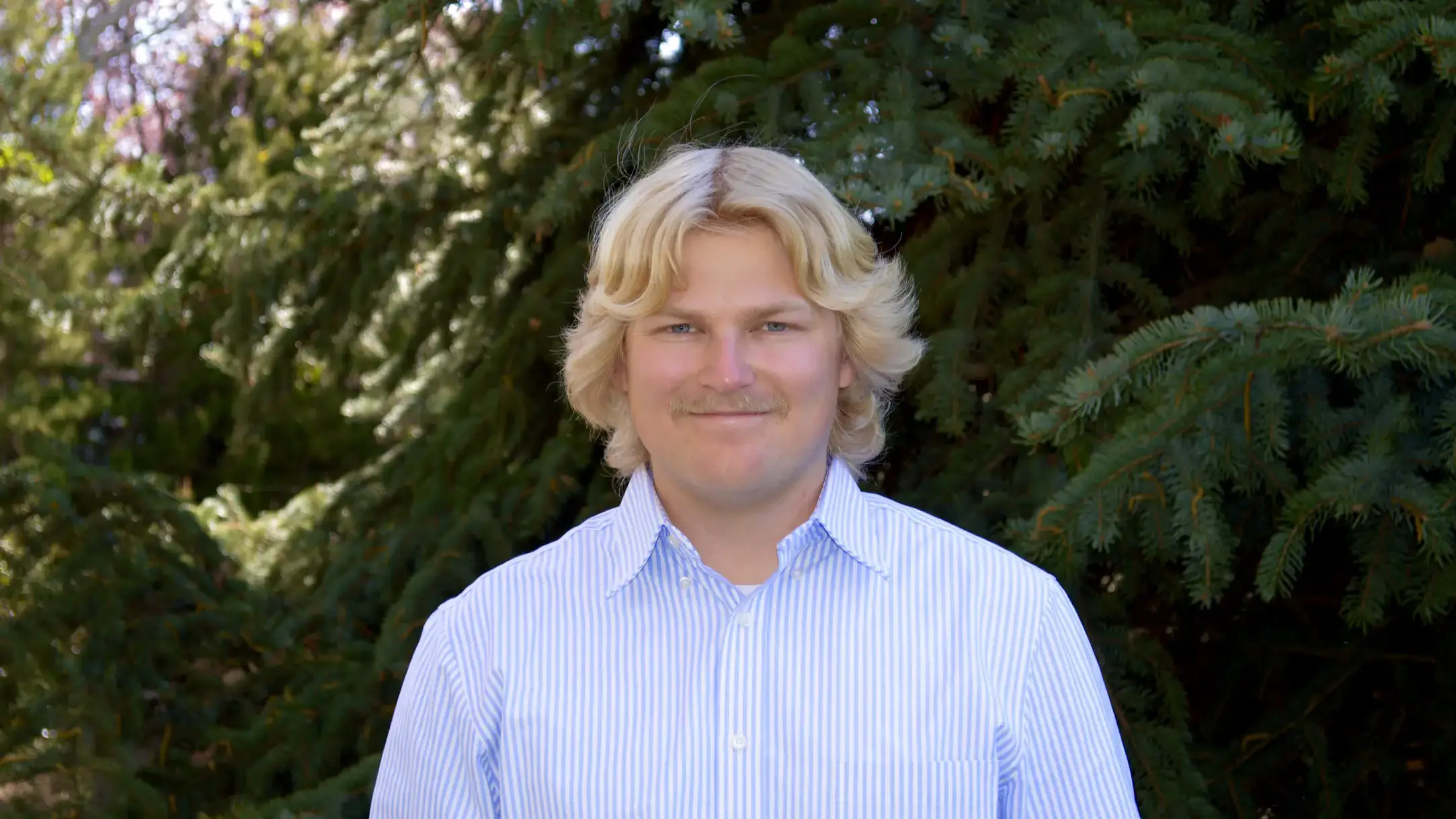 Alex Call smiles wearing a collared shirt in front of evergreen trees in background.