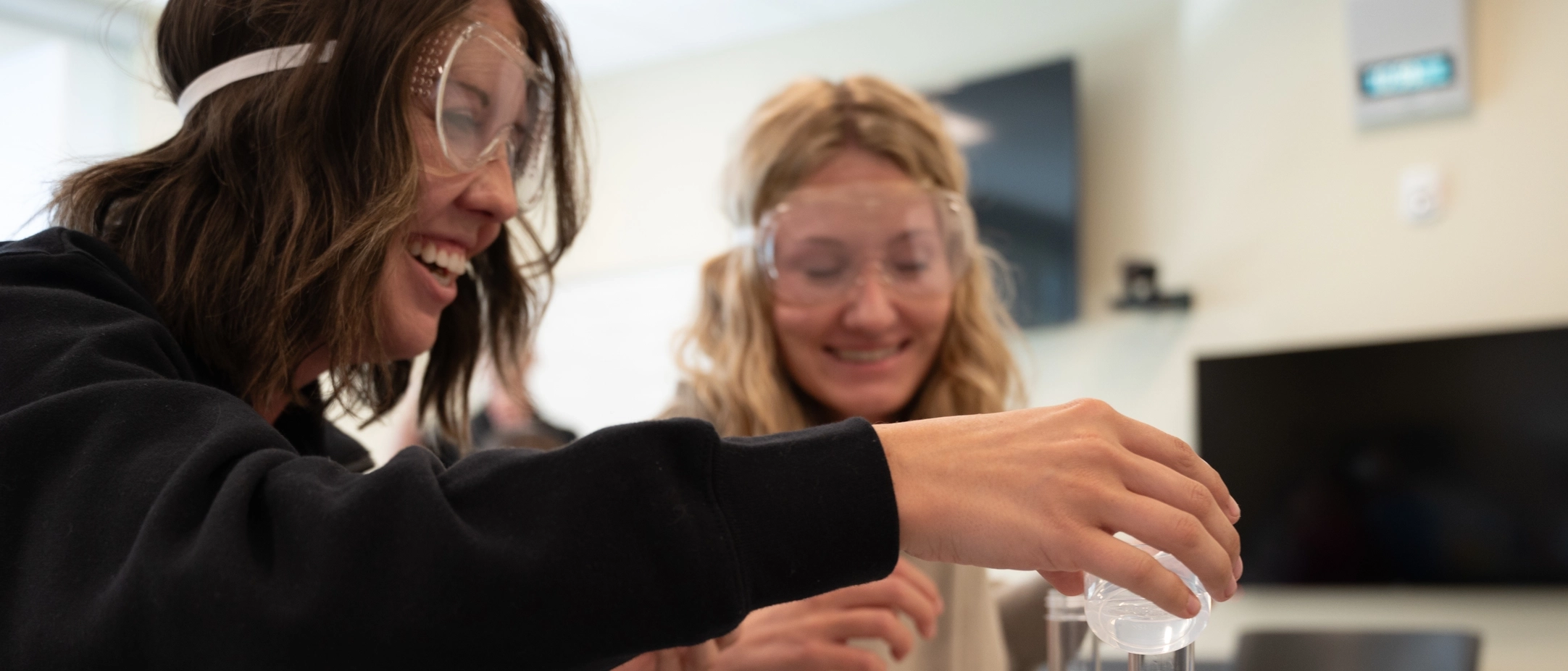 two students wearing safety goggles while one pours liquid into a clear container 