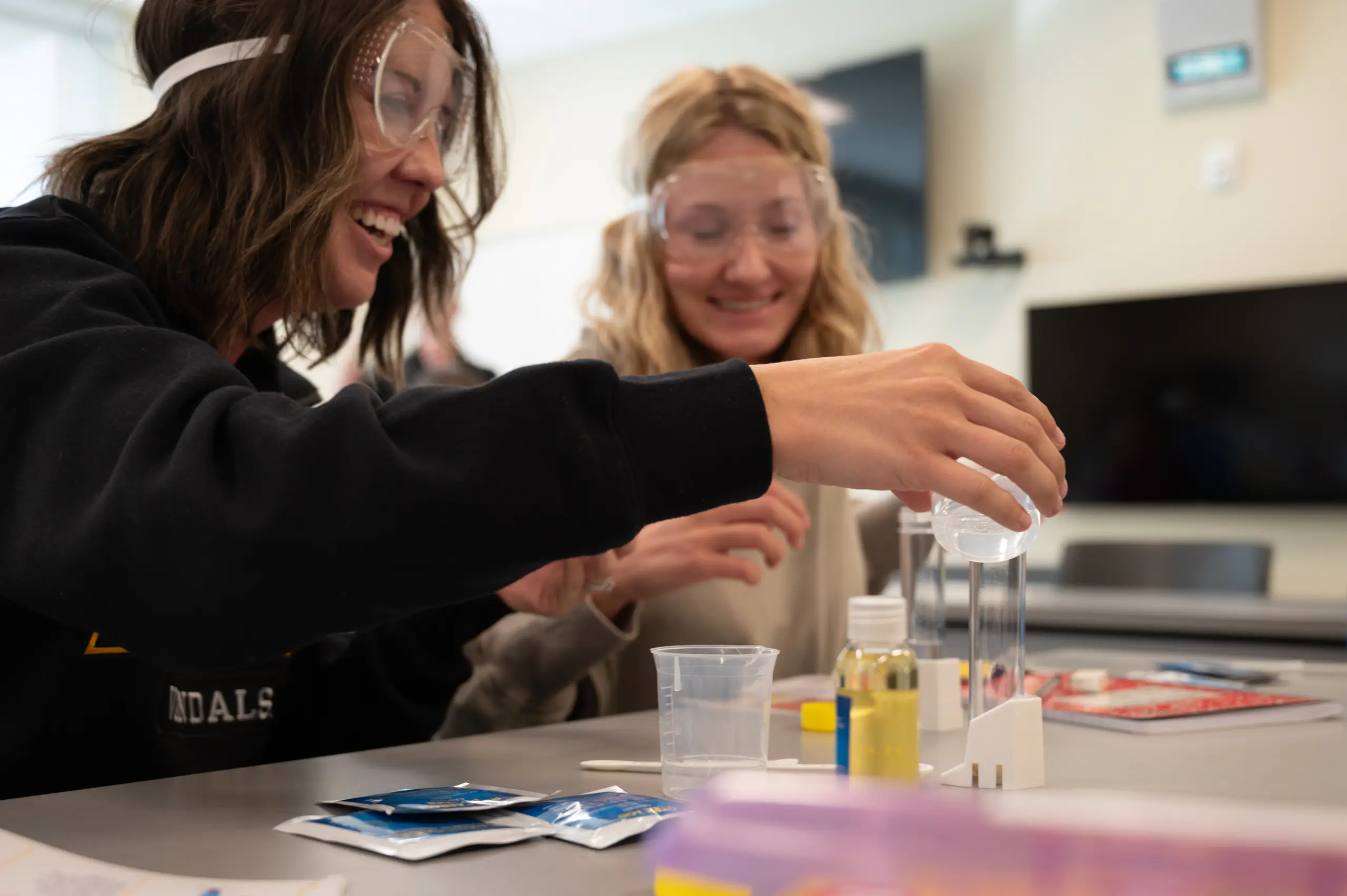 two students wearing safety goggles while one pours liquid into a clear container 