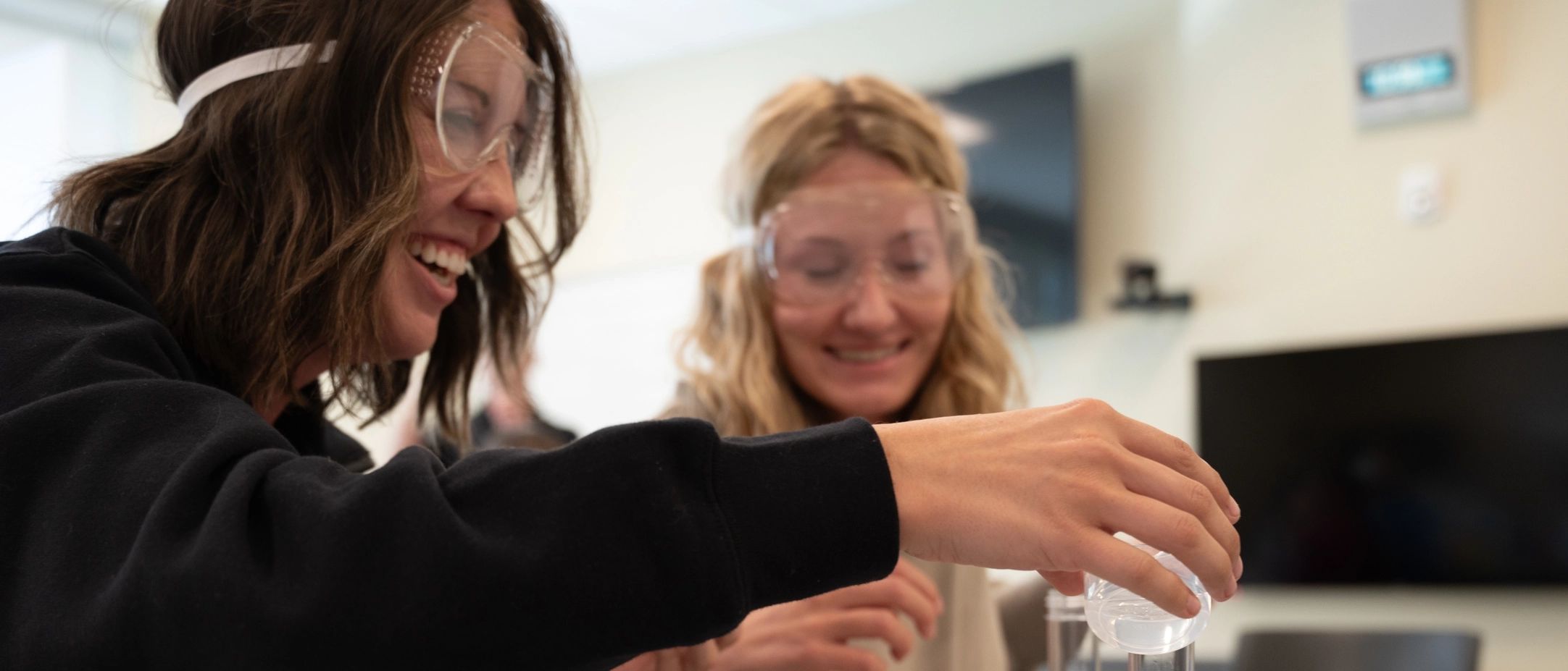 two students wearing safety goggles while one pours liquid into a clear container 