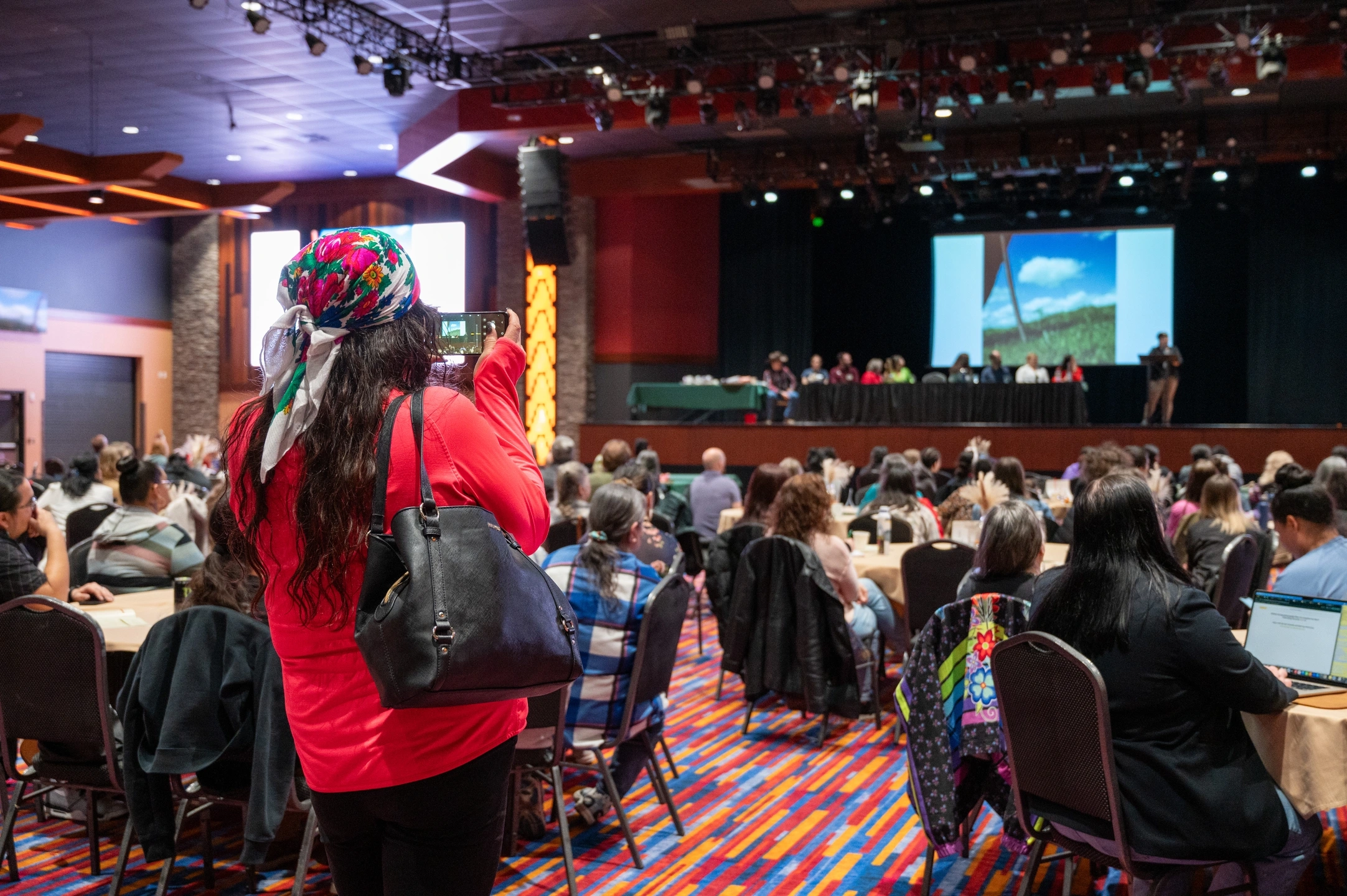 A woman takes a photo on her phone in a crowded conference room while a panelist talks on stage.