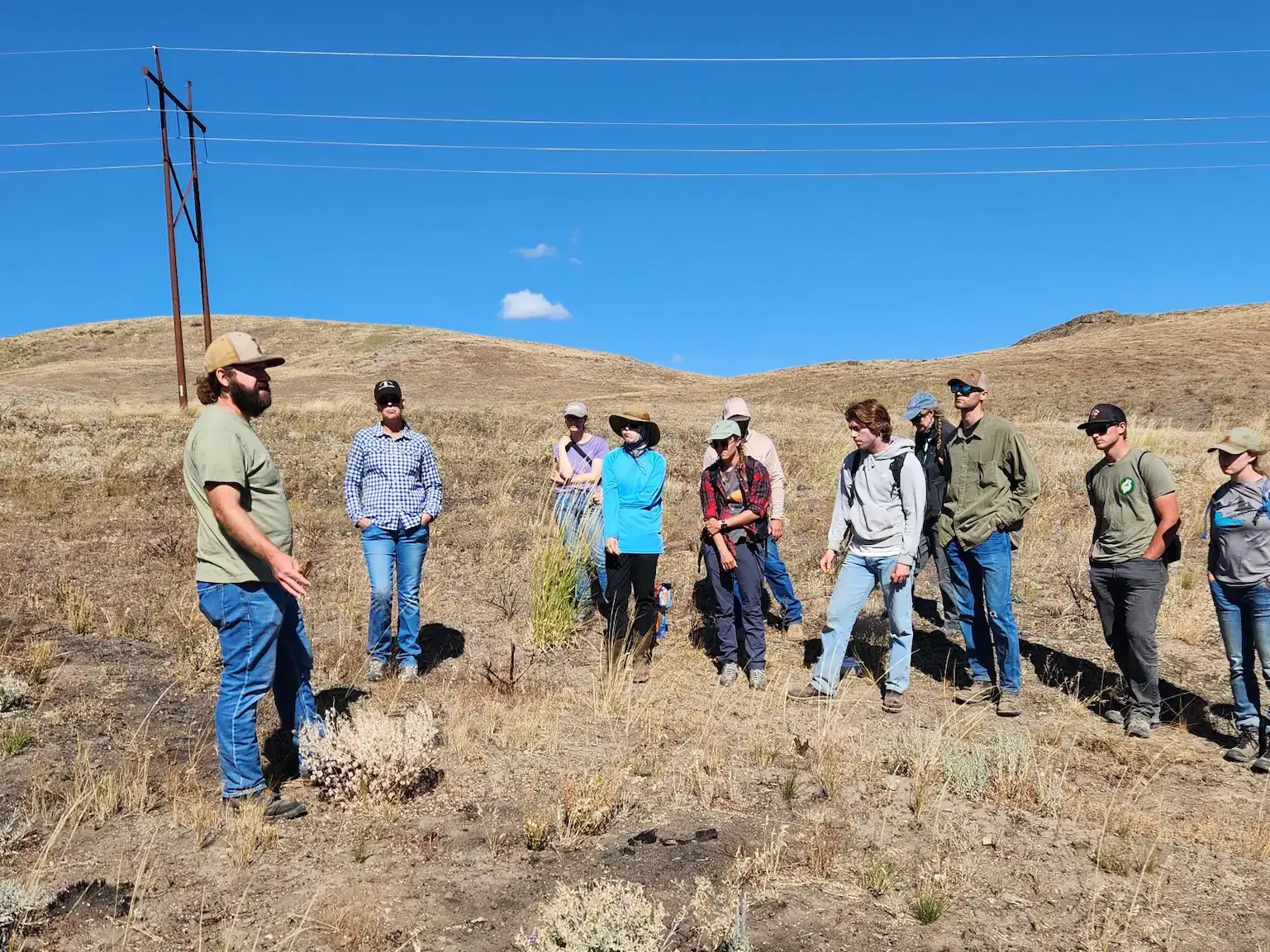 A group of students listen to a man in a tan hat give an outdoor lecture in a grassland area. 