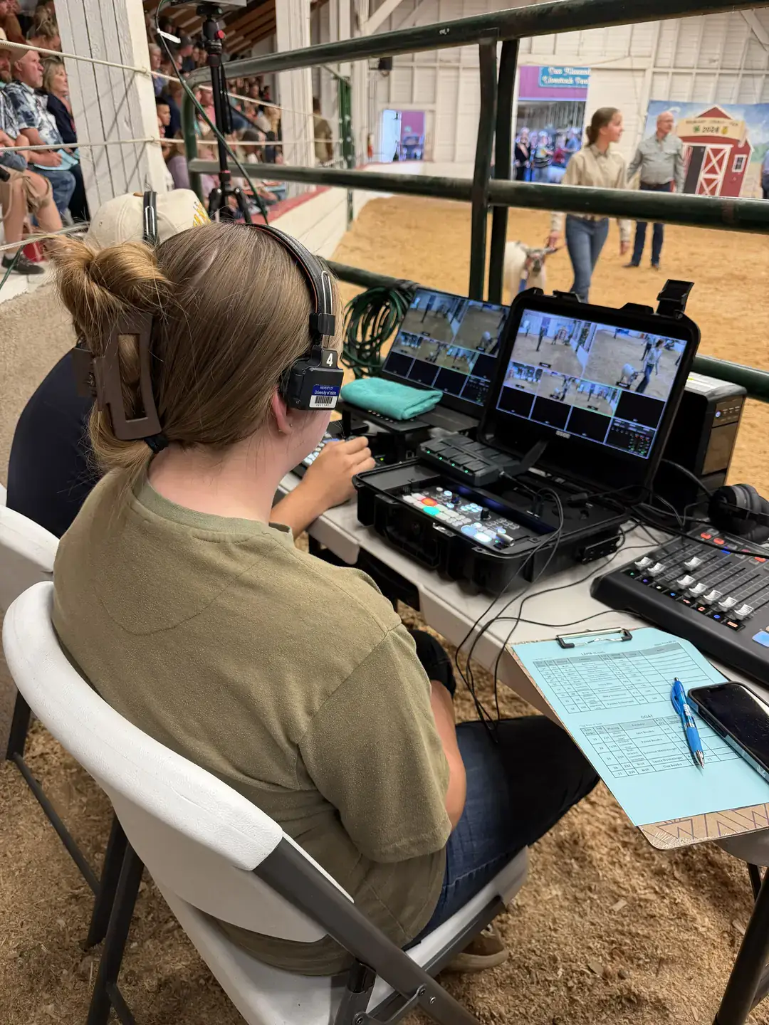 Two girls sit at a table with computer and video equipment while another girl and her goat walk through a dirt arena.