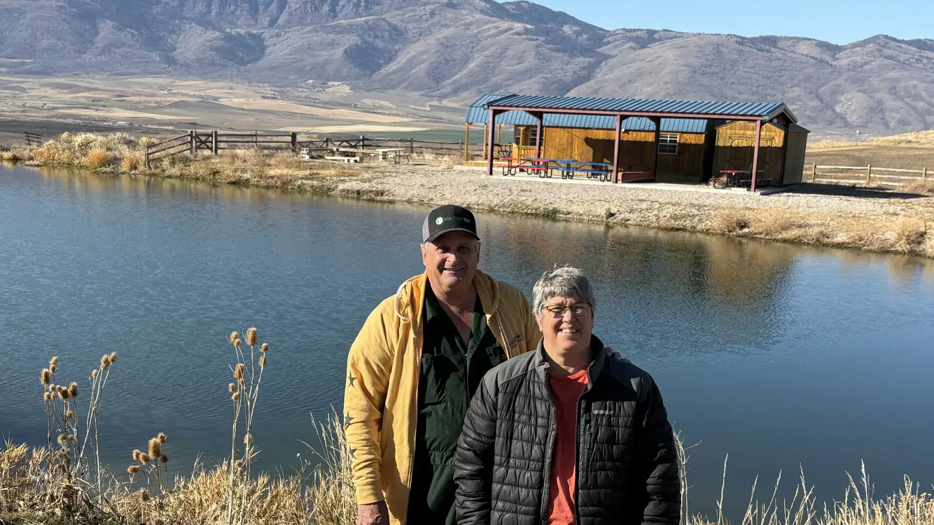 A man and a woman stand by a scenic lake with a cabin on the far bank.