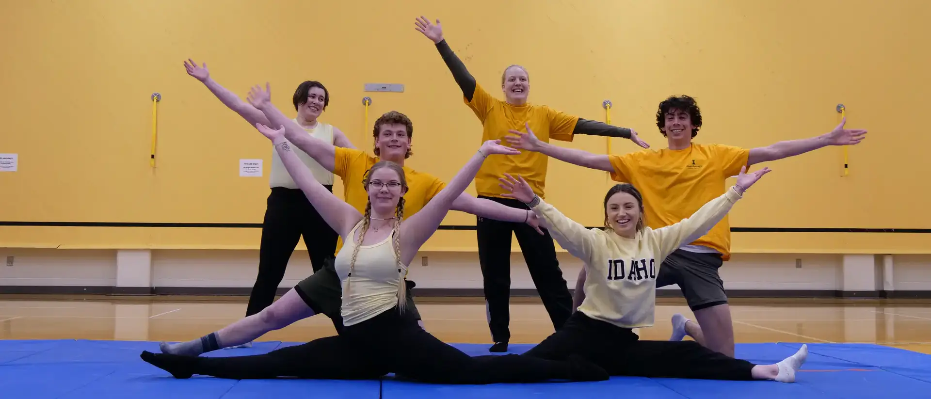 Six people posing on a gym mat, two in a splits position and four standing behind with arms extended, smiling in a gym.