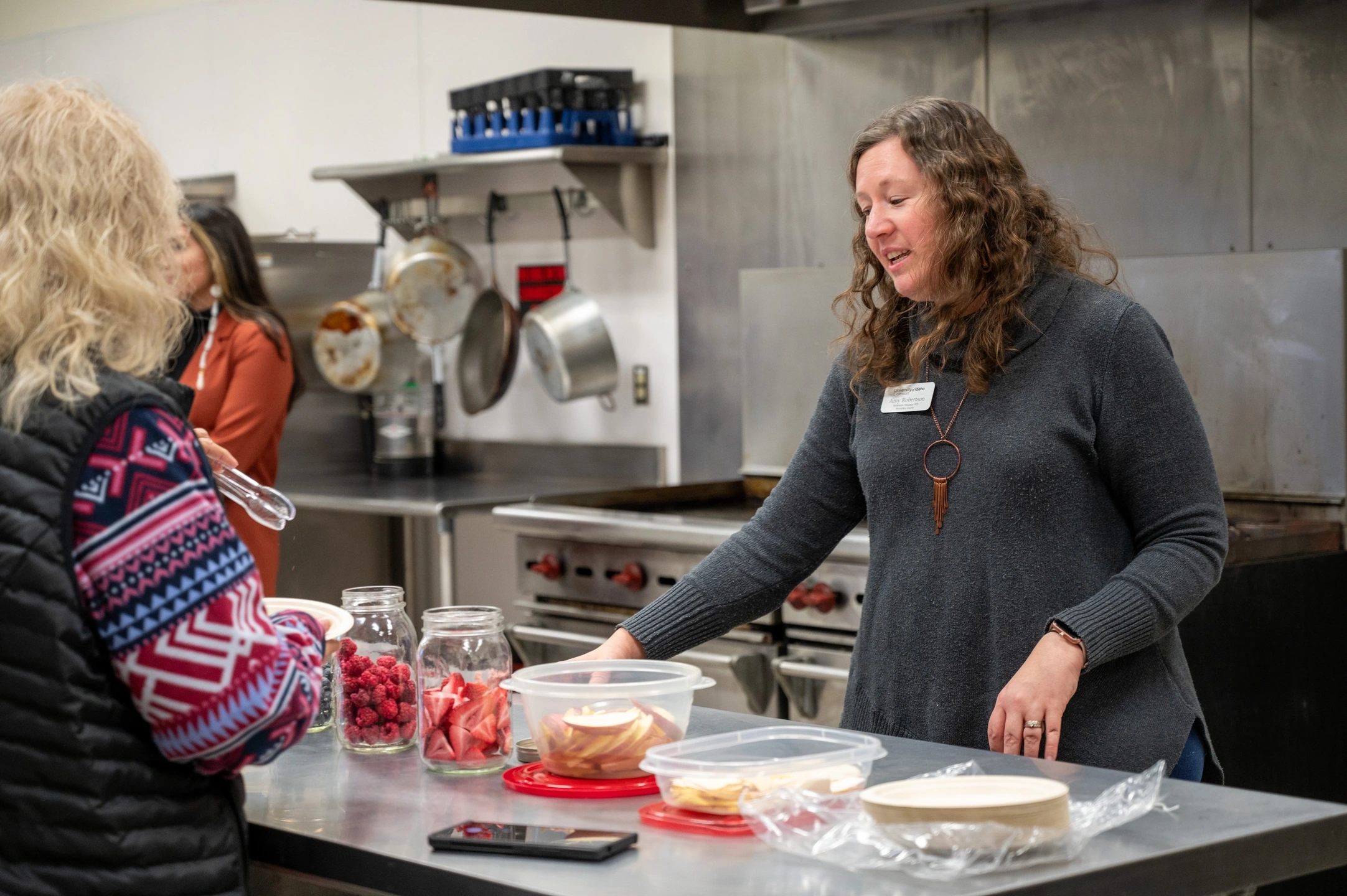 A woman gestures to a jar of dehydrated strawberries on a counter with other dehydrated fruits.