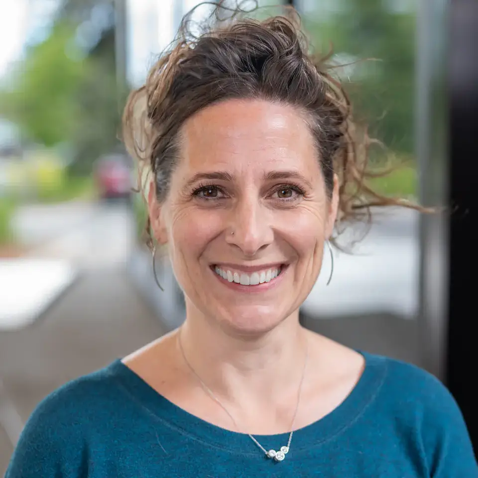 Smiling woman in a teal sweater with a necklace against an outdoor urban background.