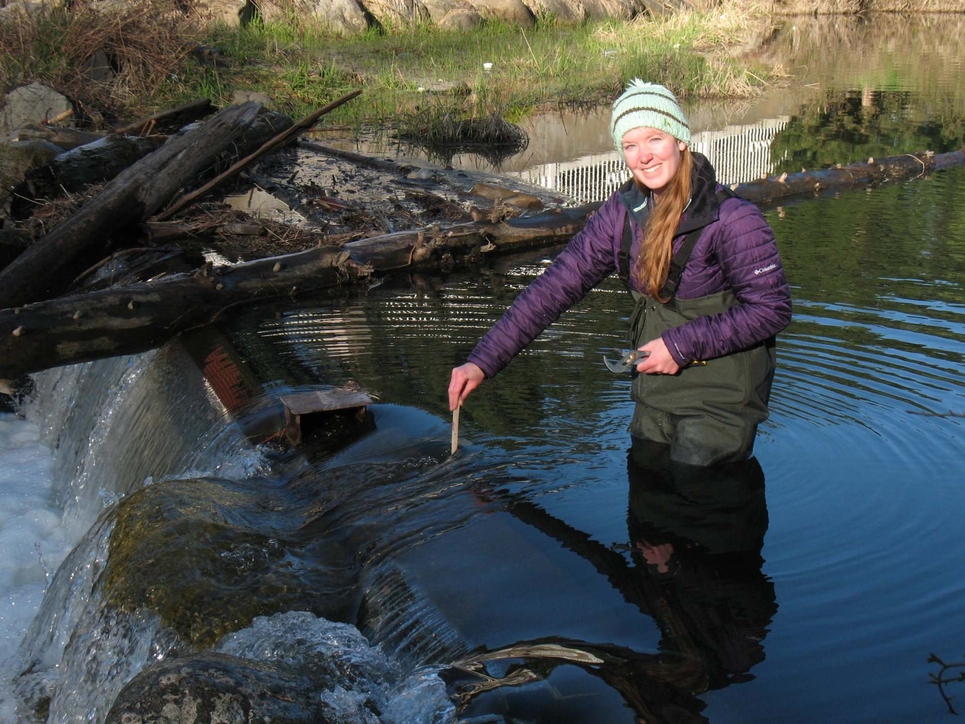 Trea LaCroix stands in a lake while wearing waders and takes measurements in an outflow of a lake.