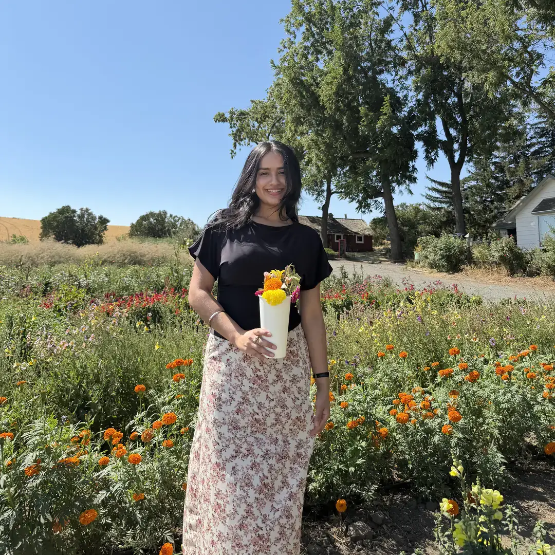 A person holding a white vase of flowers stands amidst a colorful garden with trees and a clear sky in the background.
