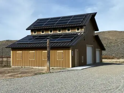 A barn with solar panels on its roof, set against a rural landscape with hills and gravel driveway.