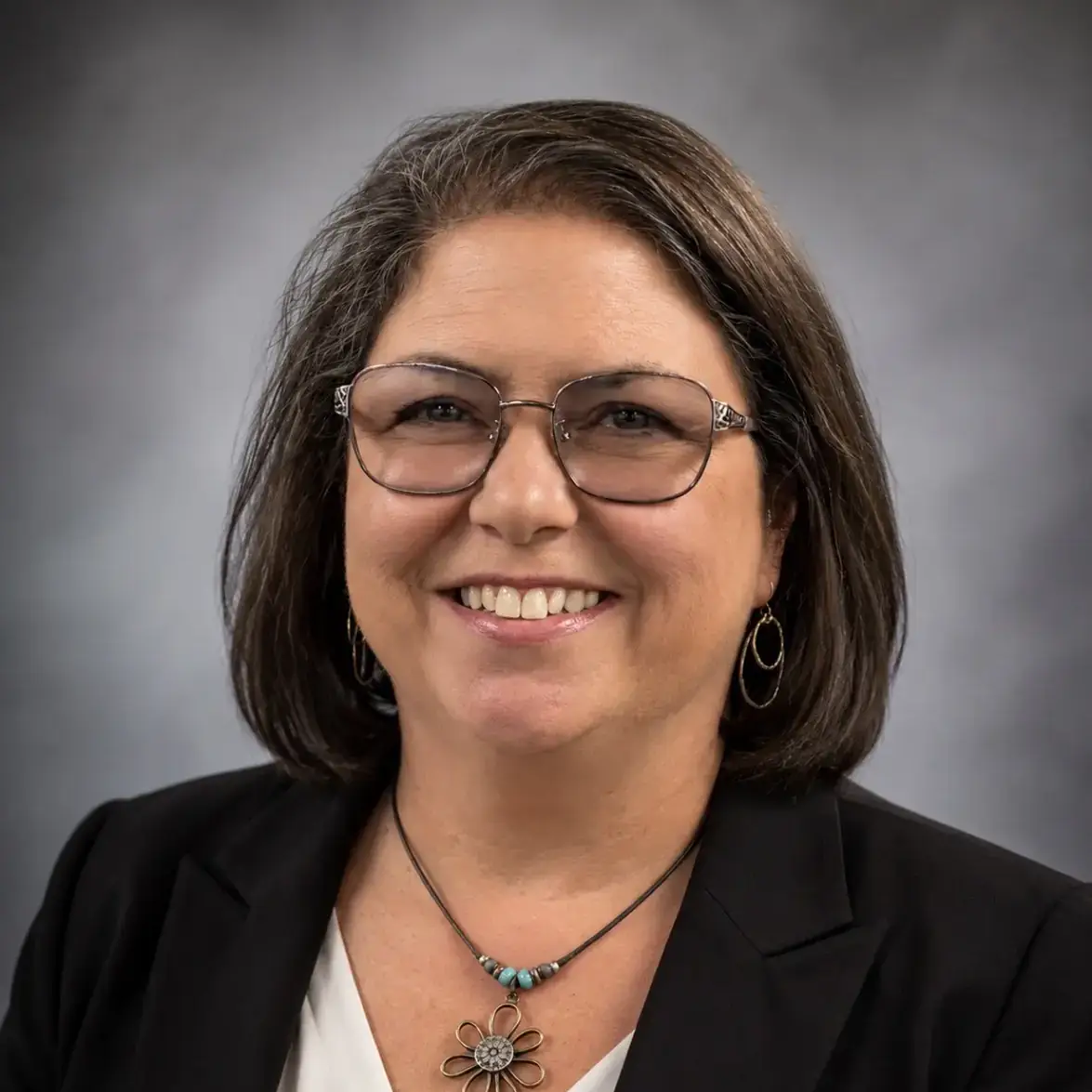 A woman wearing glasses and a black blazer, with a flower-shaped necklace, smiling in front of a gray gradient background.