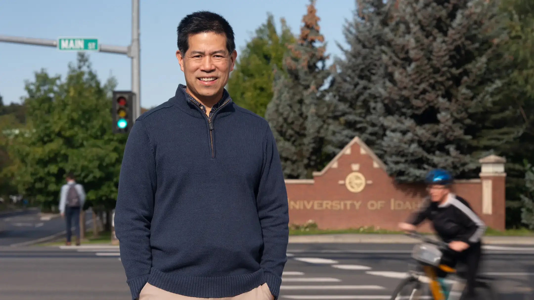 A person stands smiling in front of a University of Idaho sign, wearing a navy sweater. A cyclist and pedestrian are visible nearby.