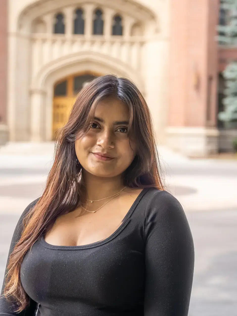 A young woman stands outdoors in front of a building with ornate architecture, smiling directly at the camera.
