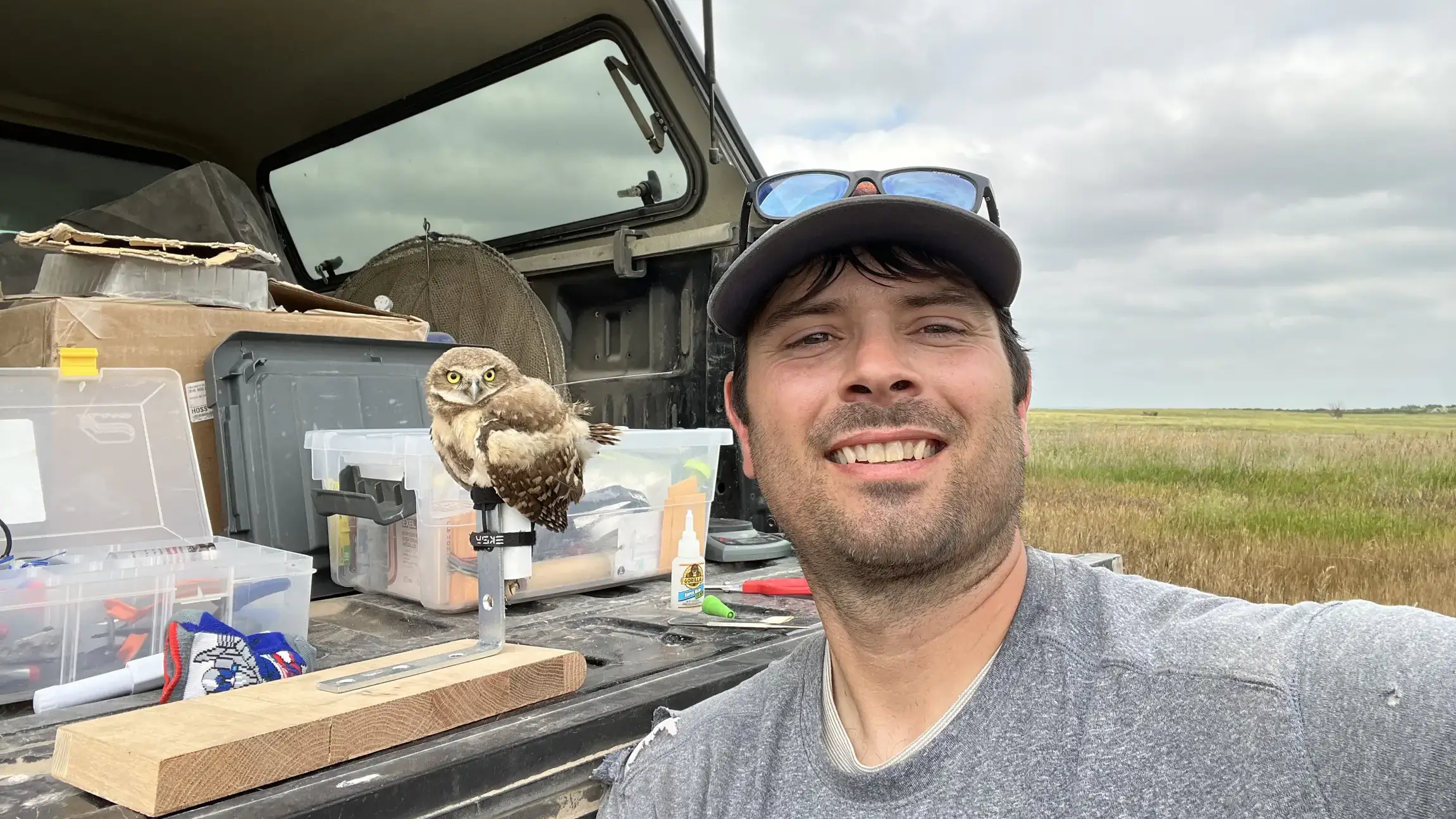 Self portrait of Anthony with pickup truck bed behind him, a burrowing owl on a small pedestal and distant prairies. 
