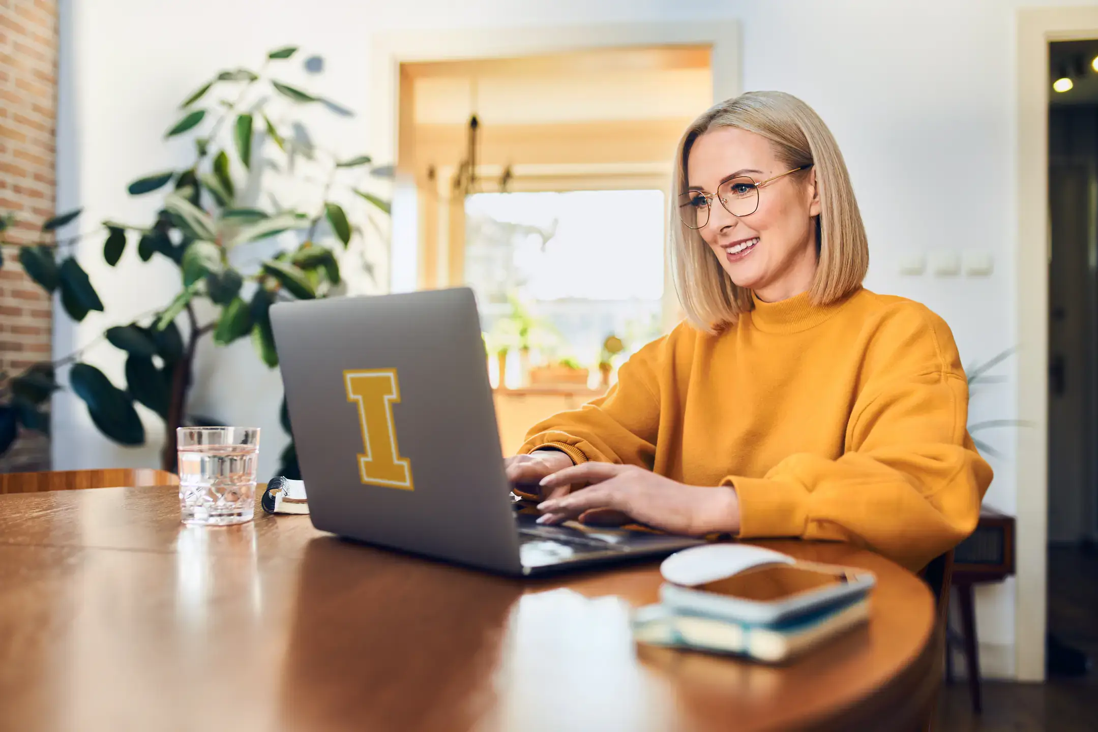 A woman wearing a gold sweater sitting at a dining table typing on a laptop computer. The computer has a vandal I shaped sticker on the lid.