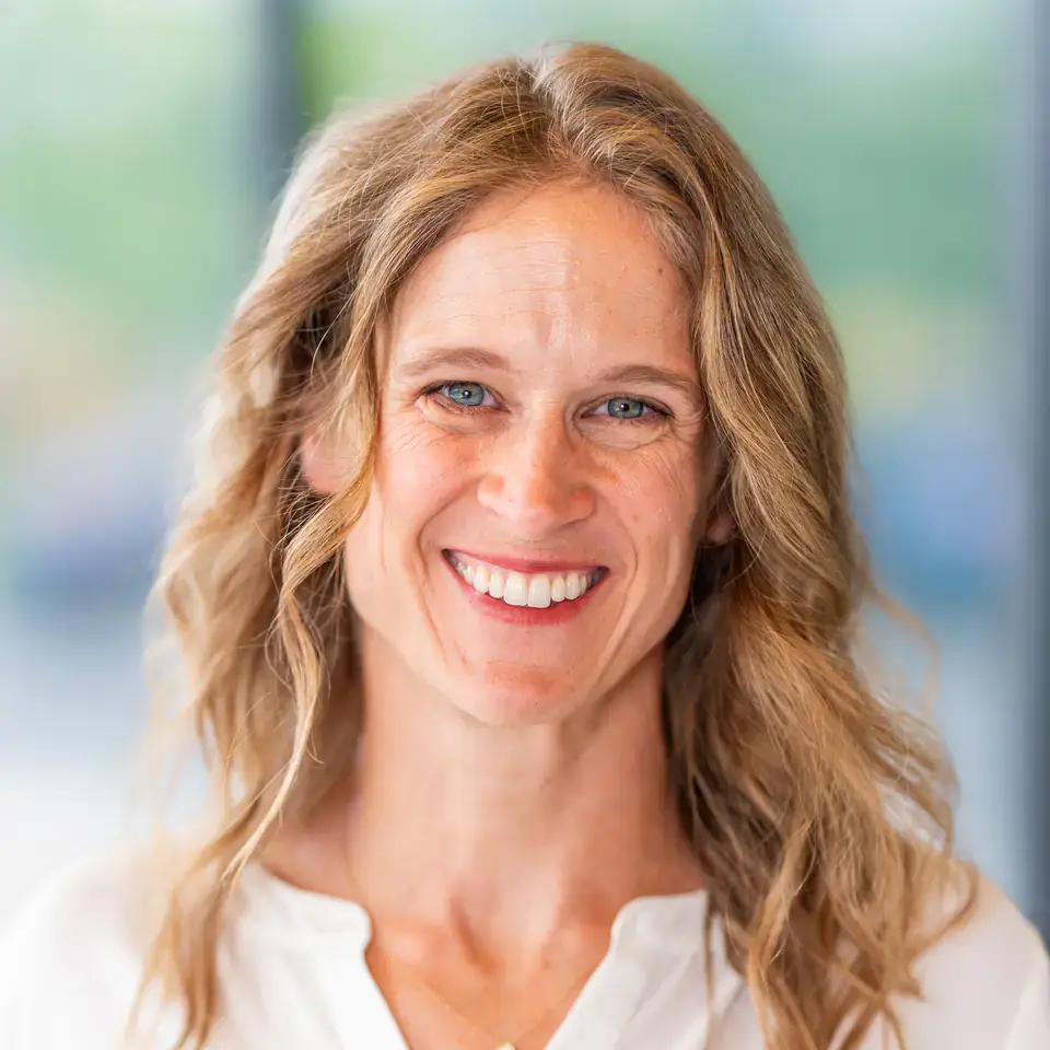 Smiling individual with wavy blonde hair wearing a white shirt, posed in front of a blurred, light-colored background.