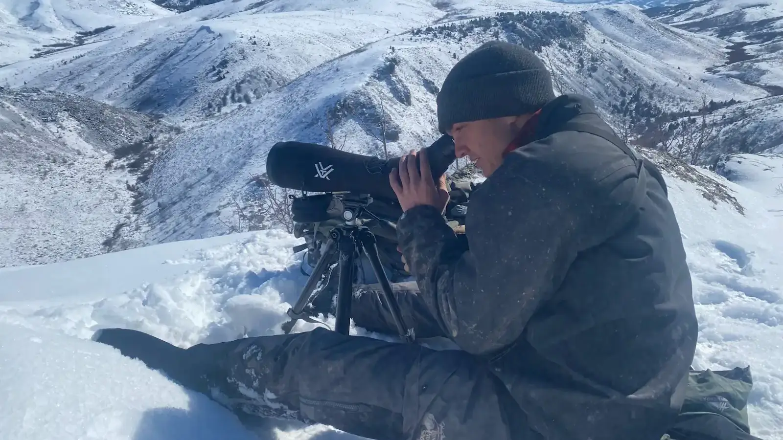 A man in an arctic setting looks through a telescope device to observe something in the distance.  