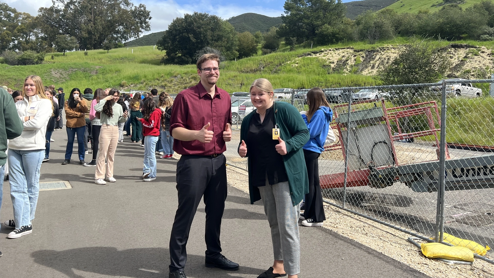 A man and women give thumbs up with high school students in the background.