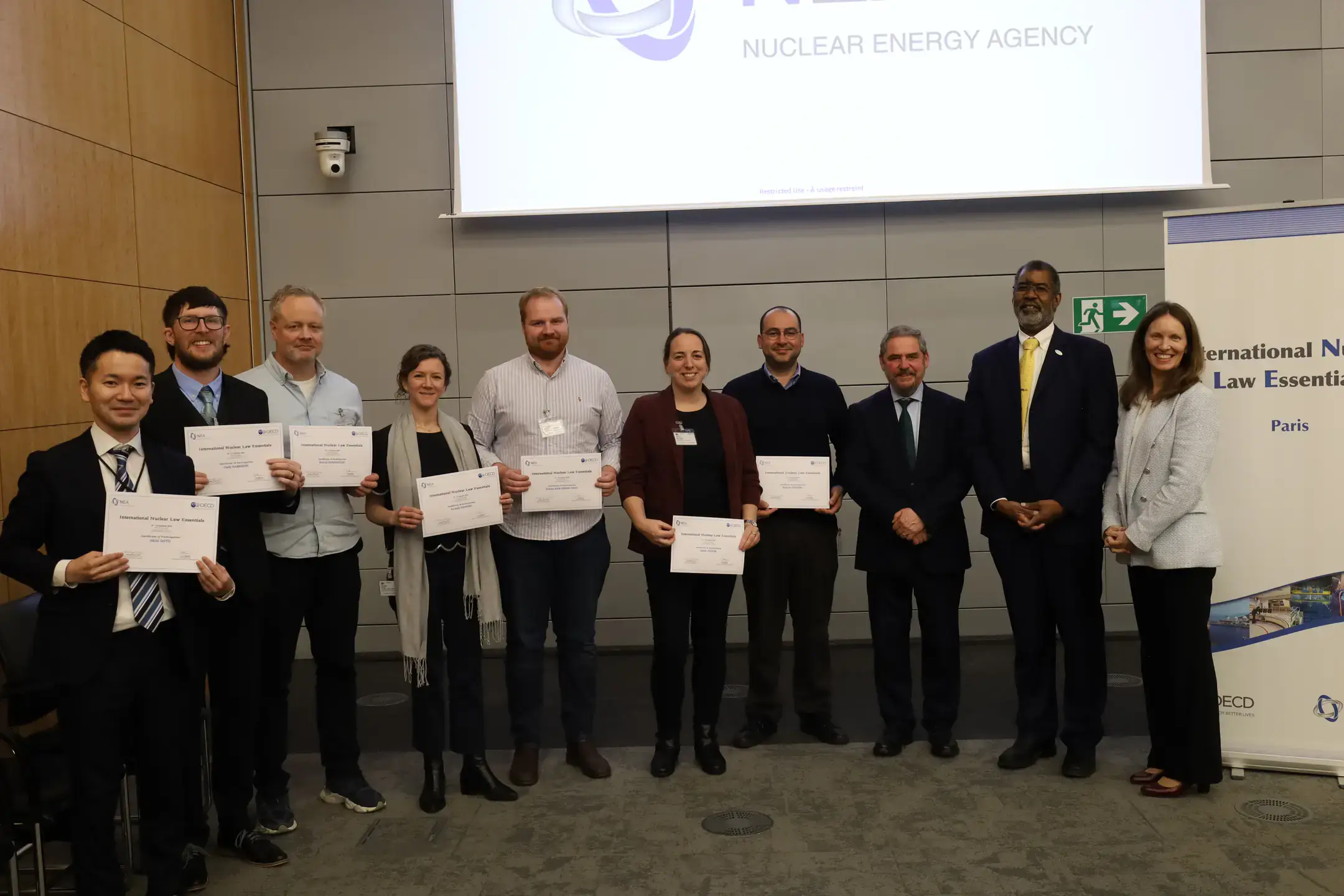 Photo of 10 men and women, seven holding certificates, in front of a banner that says nuclear energy agency.