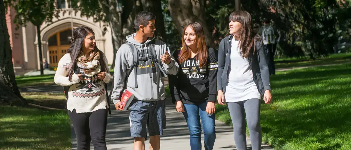 Four students walking outside on path with backpacks and books, talking and smiling