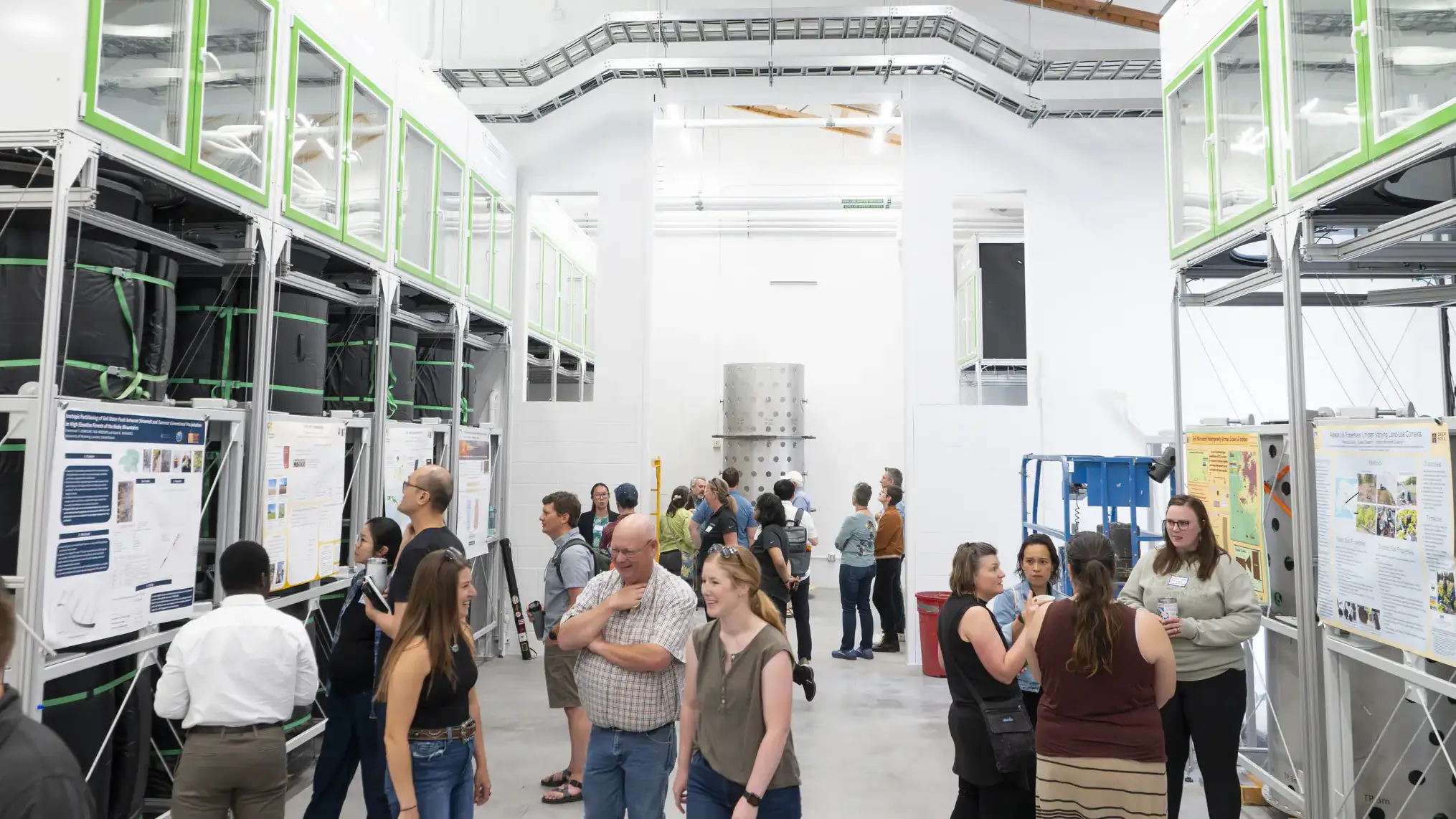 eople walk through a laboratory reading posters displayed by large, metal cylinders that hold soil.