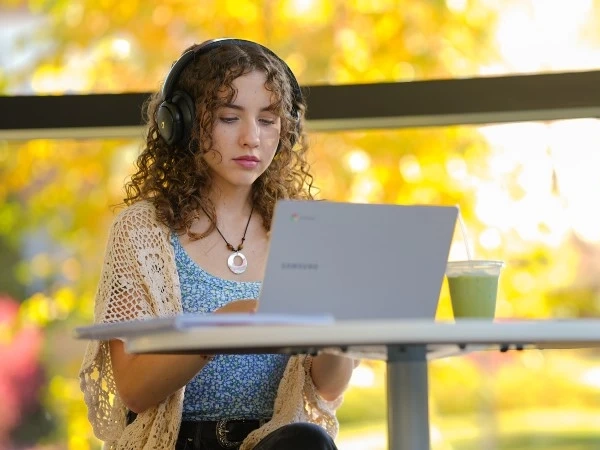 Young woman sitting at small table with laptop, view of trees in fall through window
