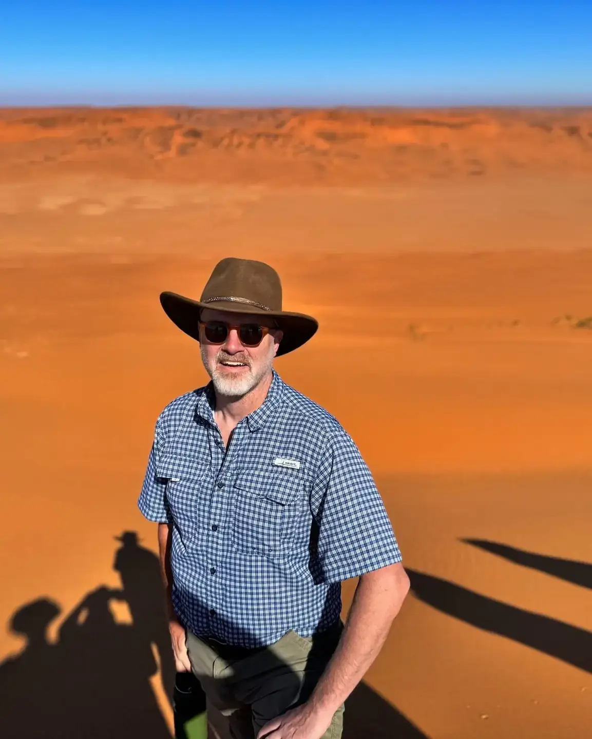 Man wearing sunglasses and a wide-brimmed hat stands on a sand dune under a clear blue sky with desert landscape in the background.