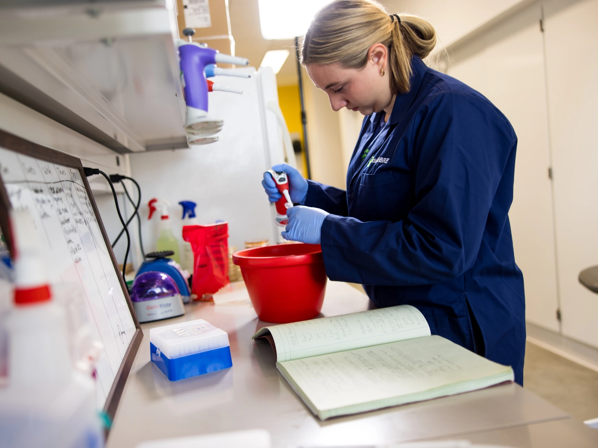 A student wearing a blue lab coat works with samples in a brightly lit lab. There’s a red boat and notebook open on the desk