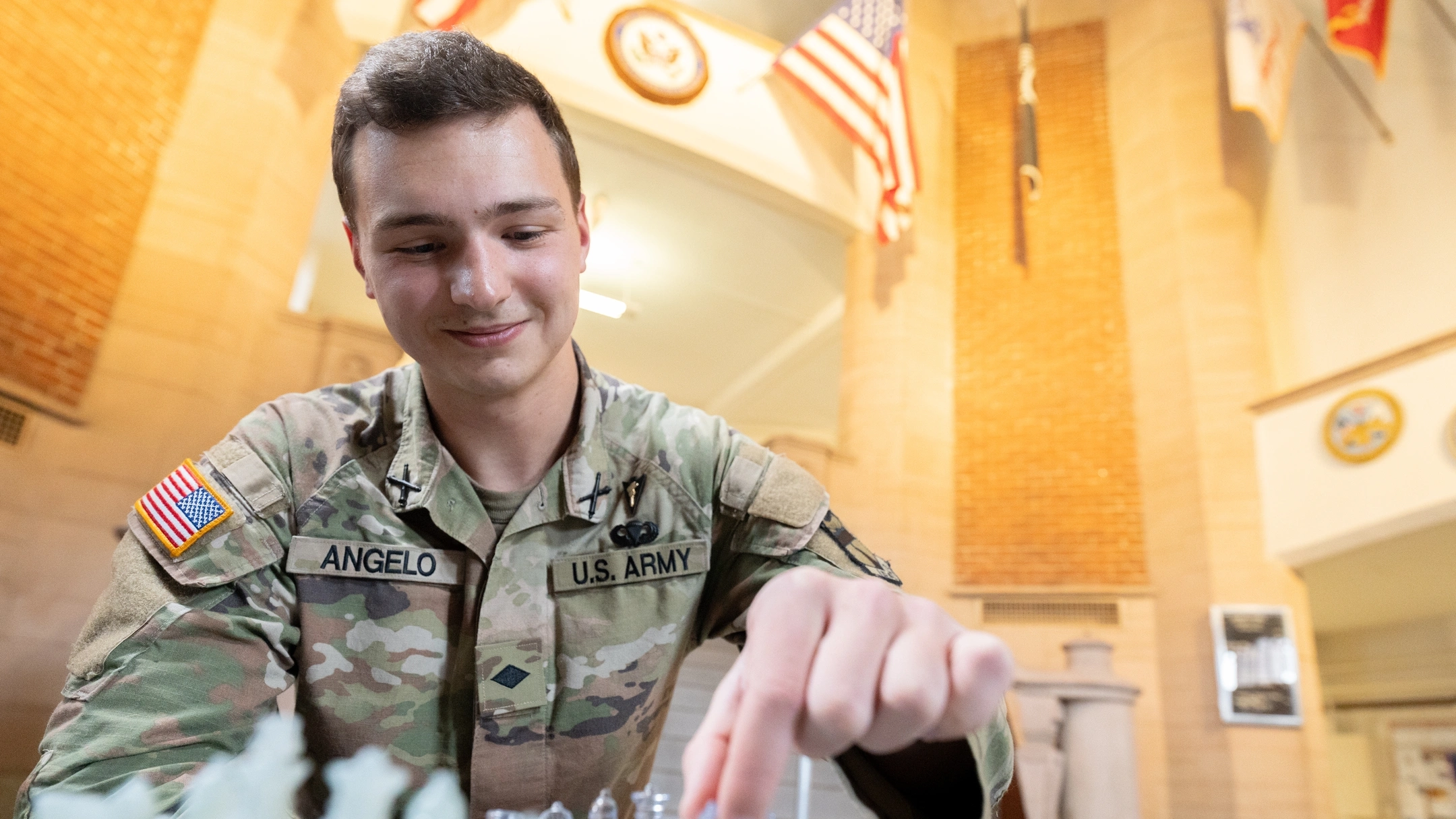 Man wearing military cammies moves a chess piece on a chess board with one hand while sitting at a table