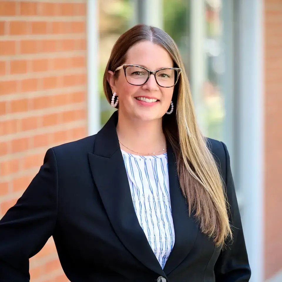 A woman in business attire, wearing glasses and smiling, stands in front of a brick wall and windows.