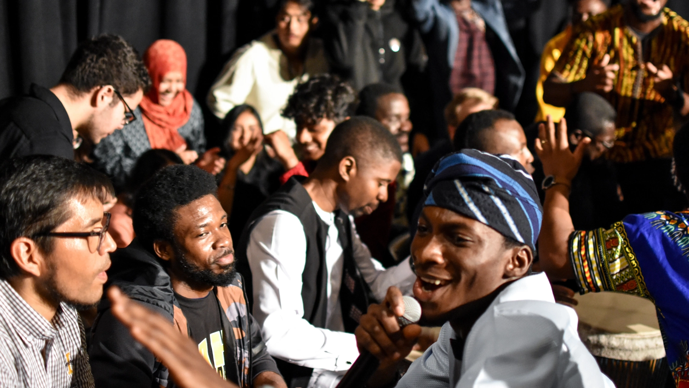 A singer amidst a group of people performance with drums visible in the background