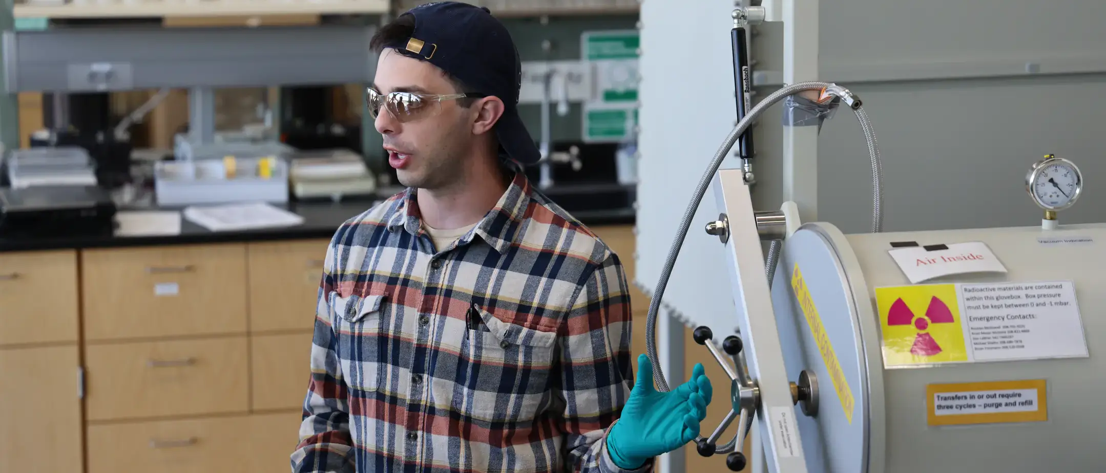 Lab worker in safety glasses and gloves speaking beside a sealed chamber with radiation warning labels