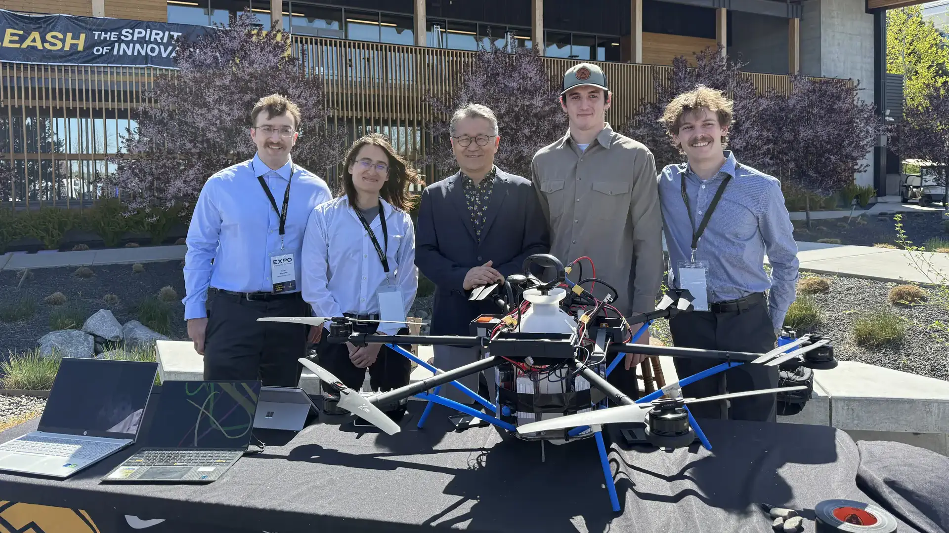 Jae Ryu stands at the center of a row of four men posing with a large multicopter drone set on a table.