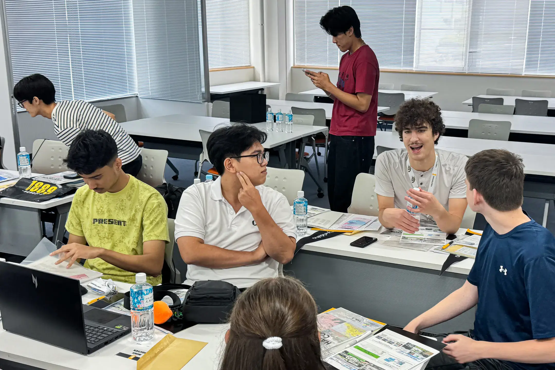 A group of people conversing and using devices in a bright classroom with desks and chairs.