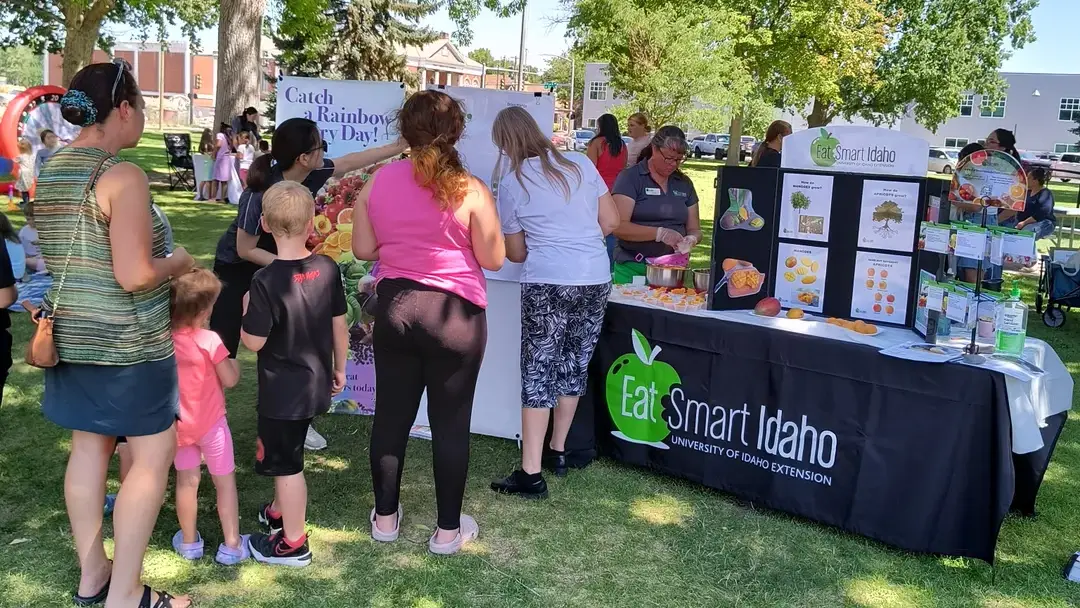 In a shady park, women and children stand in line by an Eat Smart Idaho booth for samples of various fruits in small plastic cups.