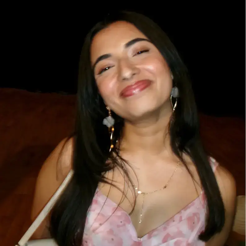 A person wearing a pink floral dress, accessorized with drop earrings and jewelry, smiling, with a dark background.
